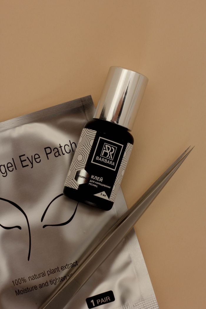 A close-up of eyelash extension tools featuring glue, tweezers, and eye patches on a beige background.