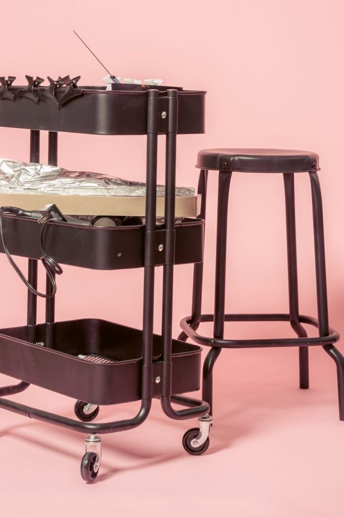 Stainless steel cart on wheels with different instruments for hairstylist near chair in studio against pink background