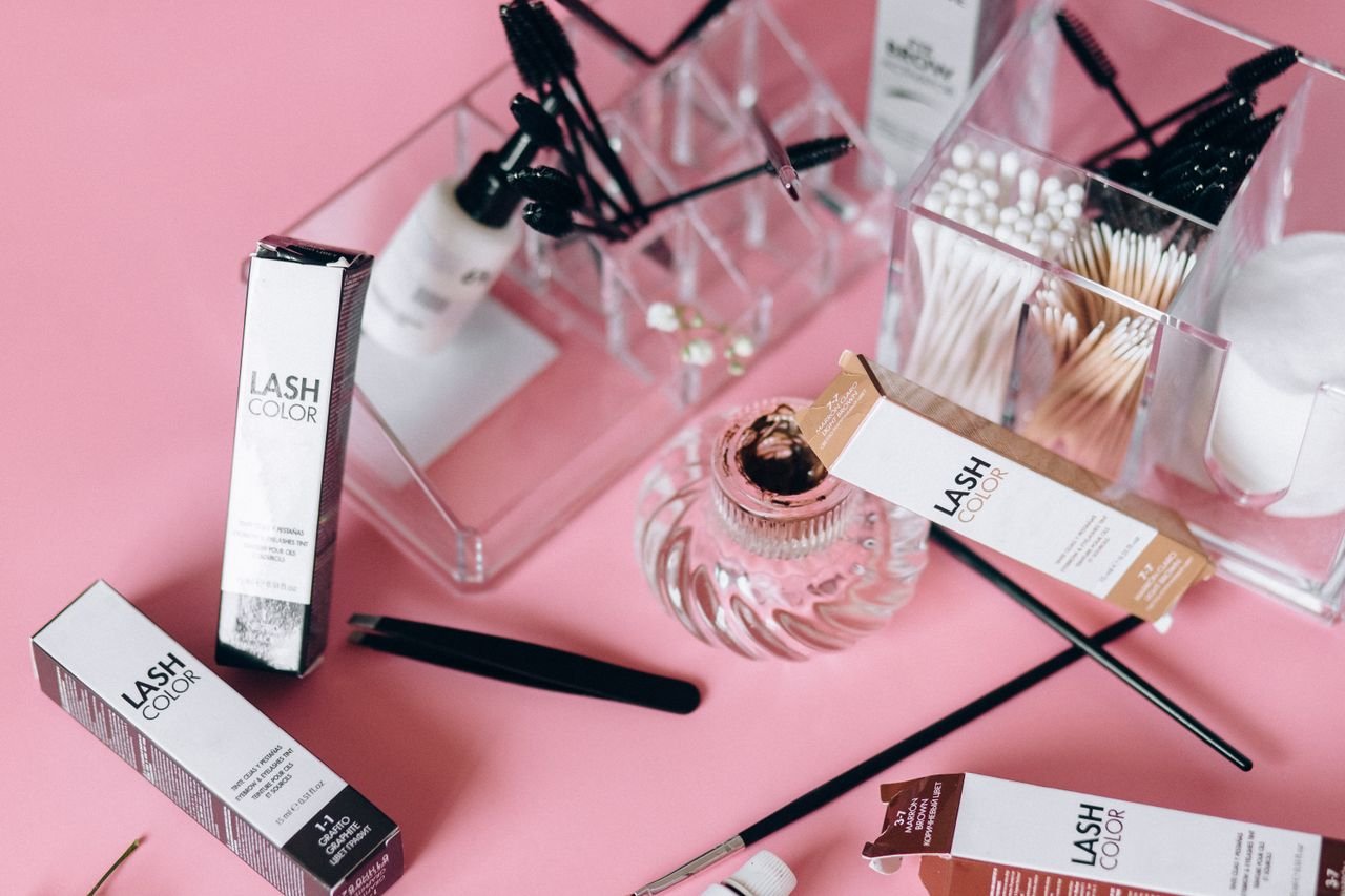 Flat lay of various cosmetic tools and products on a pink surface with clear organizers.